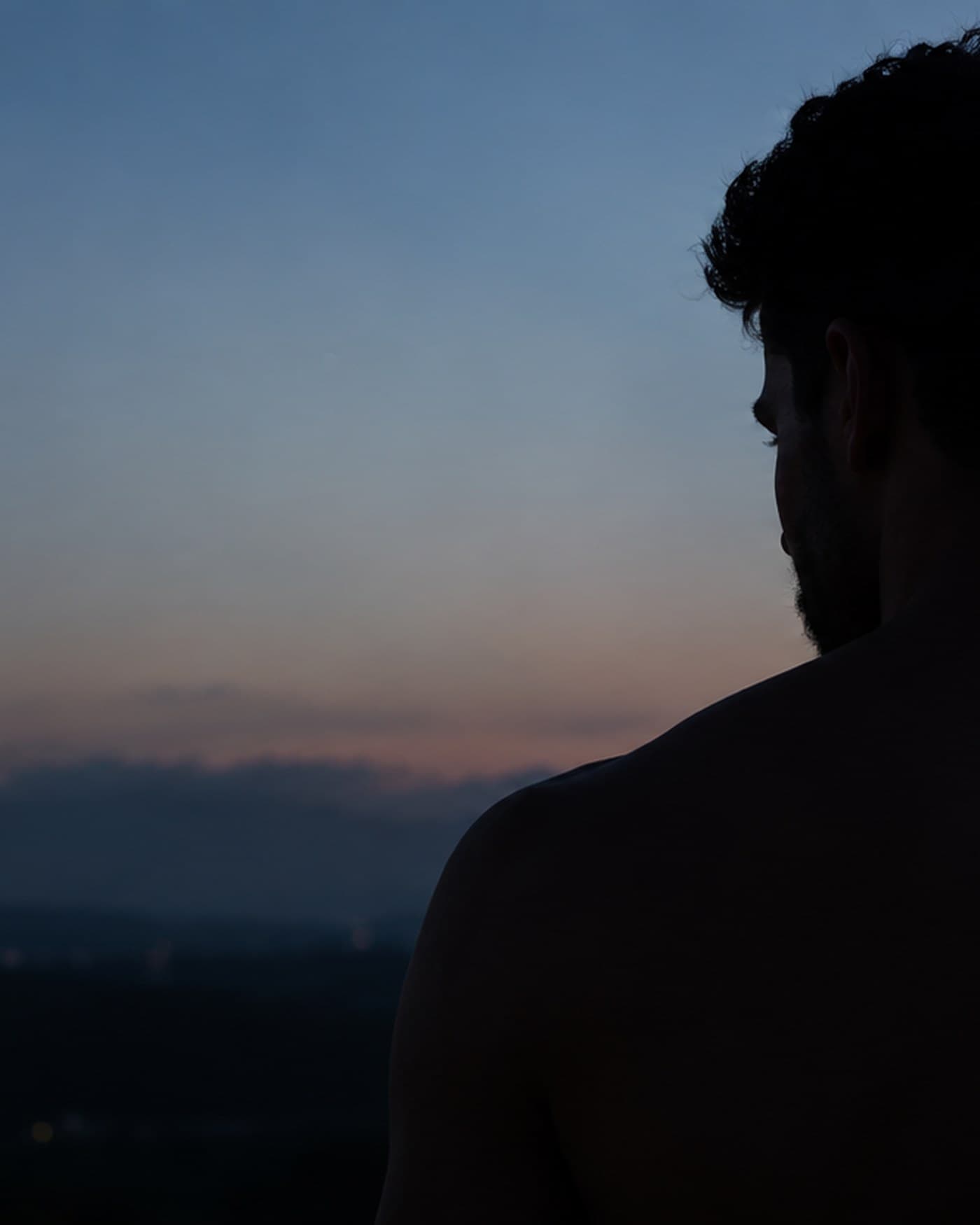 A young man alone at dusk in a hot, near-empty asphalt parking lot, hair still wet, gym bag on one shoulder, standing beside a car door he hasn't opened, desert mountains and one sodium light behind him.