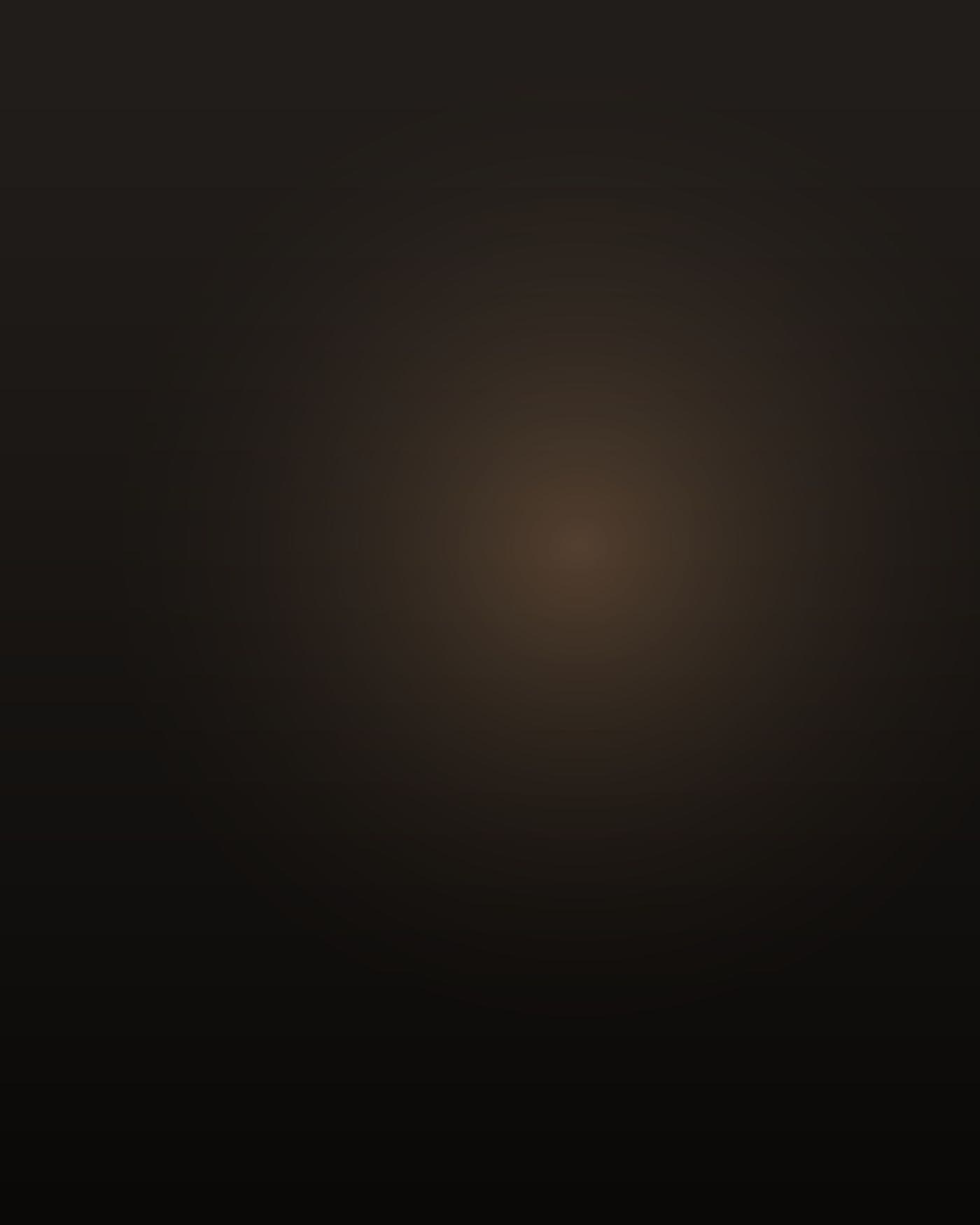 A heavyset South Asian man in his mid-thirties, Indian, thick through the chest and stomach with a heavy neck and short black hair going grey at the temples, a few days of stubble, wearing an untucked polo. He is seen from behind and slightly to the side, sitting alone in a small motel office at night, lit low and warm by a desk lamp and the glow of a television off frame. Deep shadow around him, the dark parking lot and one lit room visible through the window glass. He is not facing the camera.