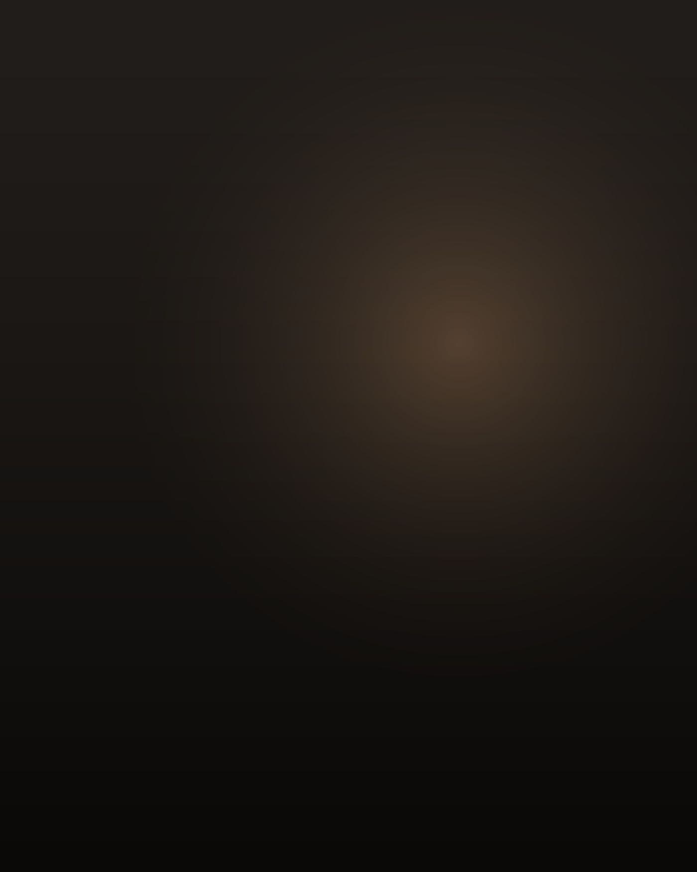 A short, thickset Filipino man in his mid-thirties, close-cropped black hair going grey at the temples, clean-shaven, heavy through the chest and shoulders, a small beige hearing aid in his left ear, standing at a dim kitchen counter before sunrise in a worn grey PT shirt, seen from behind and slightly to one side so his face is turned away, phone held against his right ear, the only light coming from the range hood over the stove and the glow of a coffee maker, unopened mail and a stack of printed pages on the counter beside him, deep shadow through the rest of the room.