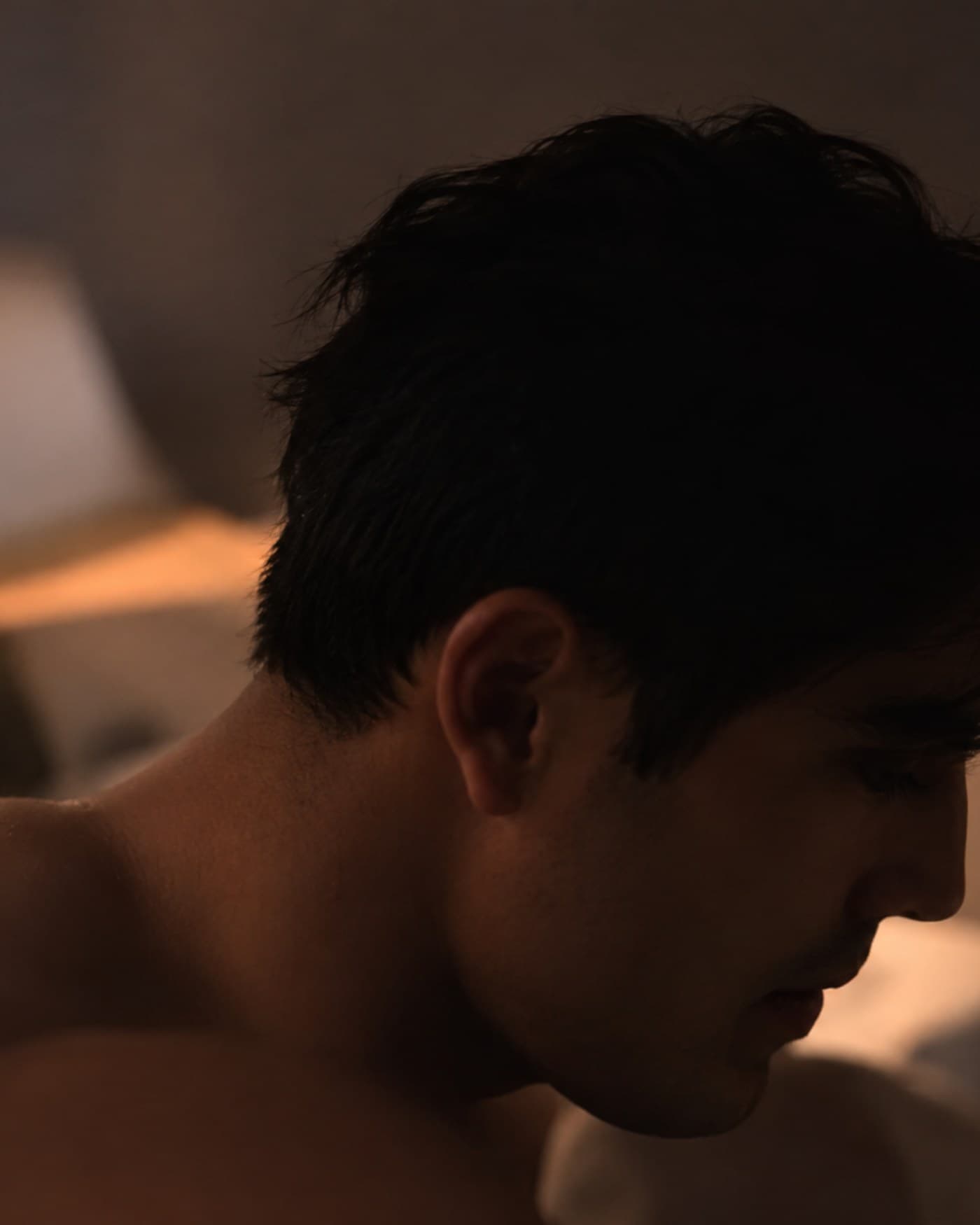 A young man alone at night in a small bedroom, one arm reaching into a half-open closet, rain on the dark window behind him, single lamp.